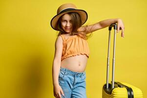 Happy child girl with stylish suitcase, cutely smiles at camera over yellow background. Travel, kids and summer concept photo