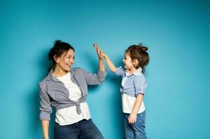 Young curly woman and cute little girl look at each other and clapping hands gesturing high five . Mother and and daughter on blue background photo