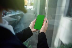 Focus on a phone with a green chroma key to insert ads in the hands of an unrecognizable woman standing in the window of a high-rise building overlooking an urban background. Copy space. photo