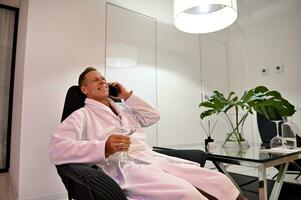 Happy attractive handsome mature Caucasian man with a drinking glass with water in hand, sitting on an armchair in wellness spa lounge zone, smiles toothy smile while talking on mobile phone photo