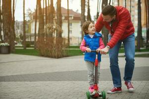 hermoso europeo amoroso y cuidando papá caminando junto a su hija montando un patada scooter mientras caminando juntos en el ciudad parque. contento familia relaciones y despreocupado infancia concepto. foto