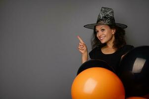 Smiling mixed race woman dressed in witch carnival costume holds colorful orange and black air balloons, points on a copy space on gray background. Concept of Halloween traditional seasonal event photo