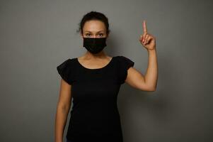 Front portrait of a cheerful African woman wearing black protective medical mask points finger up on a copy space for advertisement, isolated over gray background. Black Friday concept photo