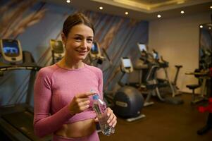 medio Envejecido bonito mujer con un botella de agua, mirando a cámara en pie a gimnasio después ejercicio. sano estilo de vida, aptitud física, deporte, culturismo, motivación, peso pérdida, cuerpo cuidado concepto. foto