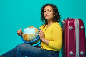 Beautiful woman in bright yellow sweater sits next to her suitcase and puts on medical protective mask on globe, isolated on blue background with copy space. Safe travel concept during pandemic. photo