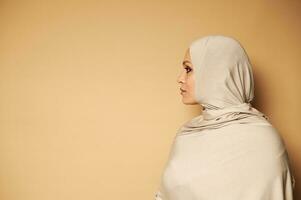 Side portrait of young Arab Muslim woman in strict religious attire and head covered in hijab looking to the side against beige background with copy space photo