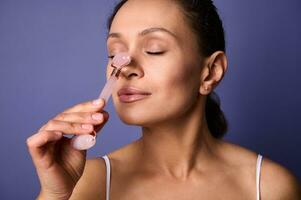 Close-up beauty portrait of middle aged pretty woman massaging her nose with stone roller massager, doing smoothing anti-aging face lifting massage with cosmetic tool, isolated on purple background photo