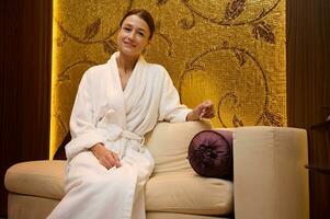 Confident portrait of a relaxed serene middle aged Caucasian pretty woman in white bathrobe sitting on a couch, smiles looking at camera while resting in a luxurious health spa lounge zone photo