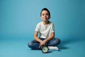 Nice boy in white t-shirt and jeans sitting on a blue background with copy space behind an alarm clock. photo