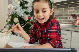 Close-up of a little vlogger, European beautiful baby girl mixing flour in glass bowl, preparing dough, talking by video call, recording video blog against a Christmas tree background at home kitchen photo