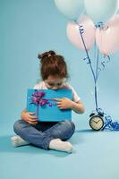 Curious child holding a gift and touching a rope of a pink bow with her finger. Alarm clock and balloons on blue background . Concept of Happy Birthday photo