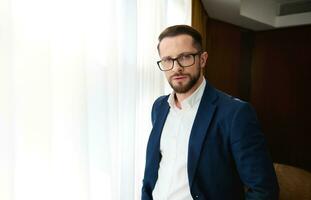 Waist-length portrait of a prosperous business investor, handsome successful young businessman standing by the window in a hotel room and looking confidently at the camera through eyeglasses photo