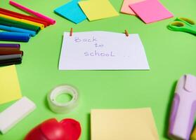 Focus on a white paper with lettering Back To School in the center of multi colored assortment of stationery office supplies and school accessories arranged in a circle. Green background, copy space photo