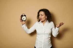 Young stupefied woman looks at an alarm clock in her hands with horror while posing on a beige background with copy space photo