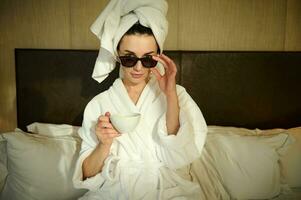 Attractive fashionable stylish young woman wearing a bathrobe, with her head wrapped in bath towel, sitting on the bed in bedroom, holding a cup of coffee and looking at camera through her sunglasses photo