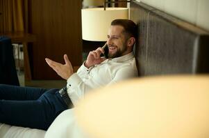 joven hermoso encantador barbado caucásico empresario vistiendo blanco camisa relajante acostado en un cama en hotel dormitorio, sonrisas mirando a cámara, hablando en célula teléfono foto