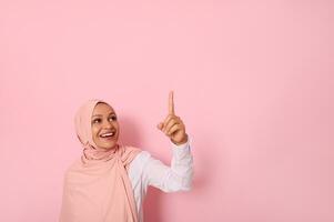 Beautiful Muslim woman of Middle Eastern ethnicity dressed in religious outfit and covered head with hijab ethnicity smiles and looks up pointing her index finger up on a copy space of pink background photo