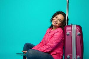 Attractive young curly haired woman with smartphone in hands sitting on the floor, leaning on her suitcase and smiles toothy smile looking at camera, isolated over blue background with copy ad space photo