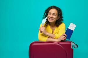 hermosa mujer en brillante amarillo suéter toma su médico máscara apagado y sonrisas con dientes sonrisa mirando a cámara, participación pasaporte con embarque aprobar, posando con maleta en azul antecedentes. Copiar espacio foto