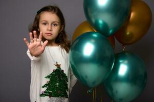 bonito niña en Navidad suéter soportes cerca hermosa brillante verde metálico y dorado aire pelotas y muestra un detener firmar, gesticulando con su dedo, aislado terminado gris antecedentes con Copiar espacio para anuncio foto