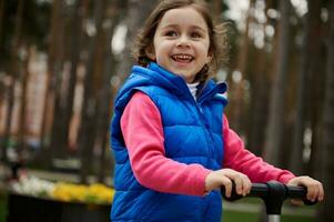 alegre sonriente contento europeo niño, linda pequeño niña paseos un patada scooter en ciudad parque. ocio actividad, contento y despreocupado infancia, activo y sano estilo de vida concepto foto