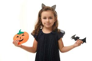Pretty little girl with a hoop in the form of cat ears, dressed in a beautiful evening dress, holds felt-cut pumpkin and handmade bat, smiles looking at camera. Isolated on white background copy space photo
