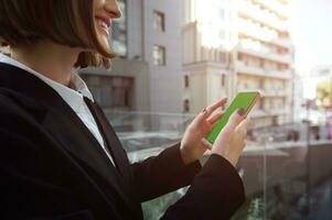 Cropped smiling brunette woman standing on balcony of high-rise building with sunbeams falling through the window, holding a smartphone with green chroma key to insert advertisement.Urban background photo