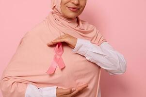 recortado vista. musulmán mujer vistiendo hijab participación rosado satín cinta a cofre nivel a espectáculo su apoyo para cáncer pacientes y sobrevivientes mundo pecho cáncer conciencia día. rosado fondo, Copiar espacio foto