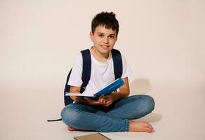 Hispanic teen boy, smart student smiles broadly at camera, holding copybook, sitting in lotus pose over white background photo