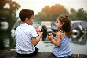 ver desde el espalda de dos niños, preescolar niña y colegio chico, comiendo sandía y mirando a cada otro, disfrutando hermosa puesta de sol sentado en el muelle a calentar verano noche foto