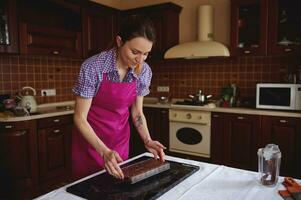 Redhead confectioner in pink apron holding chocolate molds full of liquid heated chocolate mass and putting it on a marble surface. photo