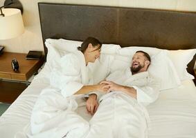 Charming couple in love of a beautiful woman and a handsome man, happy husband and wife, newlyweds, dressed in terry bathrobe, spending time relaxing in bed, enjoying the privacy of their honeymoon. photo