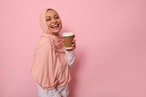 Arabic Muslim woman with covered head in pink hijab holds disposable cardboard takeaway cup, smiles toothy smile, looking at camera, standing three quarters against colored background with copy space photo