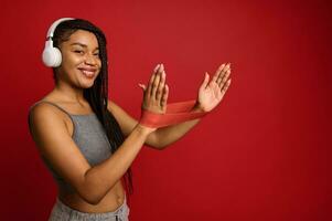 Attractive young sportswoman with stylish afro pigtails doing stretching exercises on arms with elastic fitness band, smiles toothy smile looking at camera, over colored background with copy ad space photo