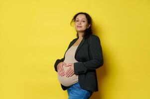 maravilloso embarazada mujer esperando bebé, formando un corazón forma con su dedos en su barriga, aislado amarillo antecedentes foto