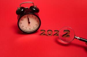 Composition with black alarm clock with midnight on the clock face and cropped hand holding magnifying glass showing number two of wooden numerals 2022. New year concept isolated over red background photo