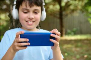 atención en moderno inteligente móvil teléfono en manos de desenfoquesonriendo Adolescente chico en auriculares, jugando en línea computadora vídeo juego foto