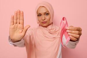 Confident portrait of blurred Arab Muslim woman in hijab holding a pink cancer awareness ribbon. Focus on woman's hand gesturing STOP , isolated over pink background with copy space. Fight Cancer Day photo