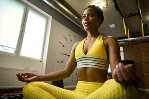hermosa africano americano mujer con plano barriga y sano cuerpo, vistiendo brillante amarillo chandal sentado en loto posición y meditando yoga, relajación ejercicios y activo vida estilo concepte foto