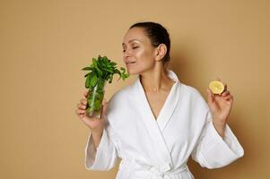 Isolated portrait on beige background of a young beautiful woman with clean skin holding a half lemon in one hand and enjoying the scent of mint in the other hand. photo