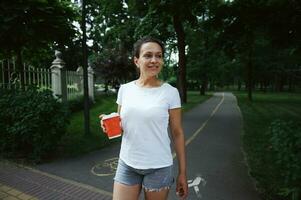 hermosa africano americano joven mujer participación para llevar café en alacena taza, caminando a lo largo el callejón de un ciudad parque foto