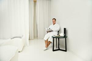 Handsome mature European business man wearing white terry bathrobe, sitting on an armchair and reading magazine while relaxing in privacy spa room of wellness spa center after hard work photo