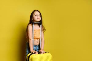 contento niño niña en elegante brillante verano ropa, con amarillo valija, mirando a cámara, aislado en amarillo antecedentes foto
