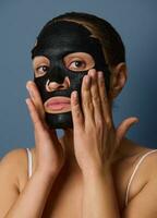 Close-up portrait of beautiful woman looking at the camera while putting on a nourishing smoothing moisturizing black face mask taking care of the beauty of her skin. Isolated on blue-gray background photo