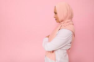 confidente de cerca retrato de árabe musulmán maravilloso mujer con atractivo mirada , vistiendo un rosado hijab mirando con confianza en frente de sí misma en pie oblicuo en de colores antecedentes con Copiar espacio foto