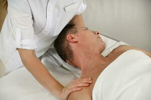 Top view of a mature man wrapped in white terry towel and lying on a massage table, getting shoulders massage in luxury wellness spa center. Alternative therapy and professional body care concept photo