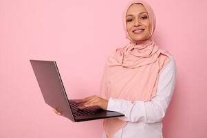 Business portrait of a successful programmer Muslim woman in pink hijab standing against a colored background with an open laptop in hands and typing text, smiling looking at camera, copy space photo