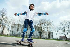 contento y sonriente chico en protector engranaje y casco mantiene equilibrar mientras montando un patineta foto