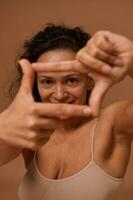 Close-up of a cheerful dark-haired curly woman wearing underclothing, smiles looking at camera through a finger frame, isolated over beige background with copy space for ad photo