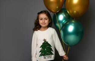 Portrait on gray background with copy space for New Year advertising of a beautiful little girl in Christmas white sweater, holding green metallic and gold air balls, cutely smiling looking at camera photo